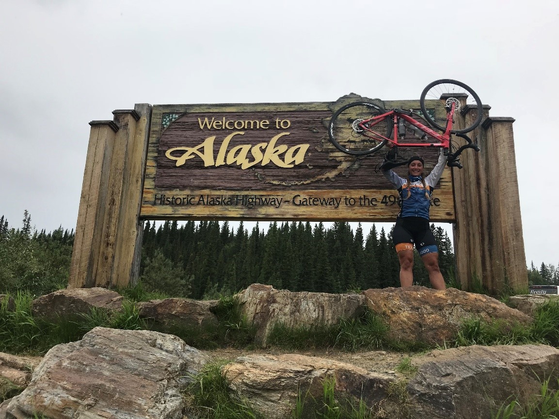 Edberg holding bike over her head infront of a welcome to Alaska sign